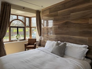 Un dormitorio con una cama grande, una silla, una ventana con cortinas y vistas a la vegetación exterior.