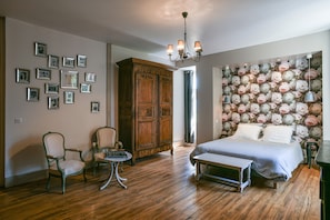 A bedroom with a bed, a wooden wardrobe, a chair, a small table, and a wall decorated with floral wallpaper.