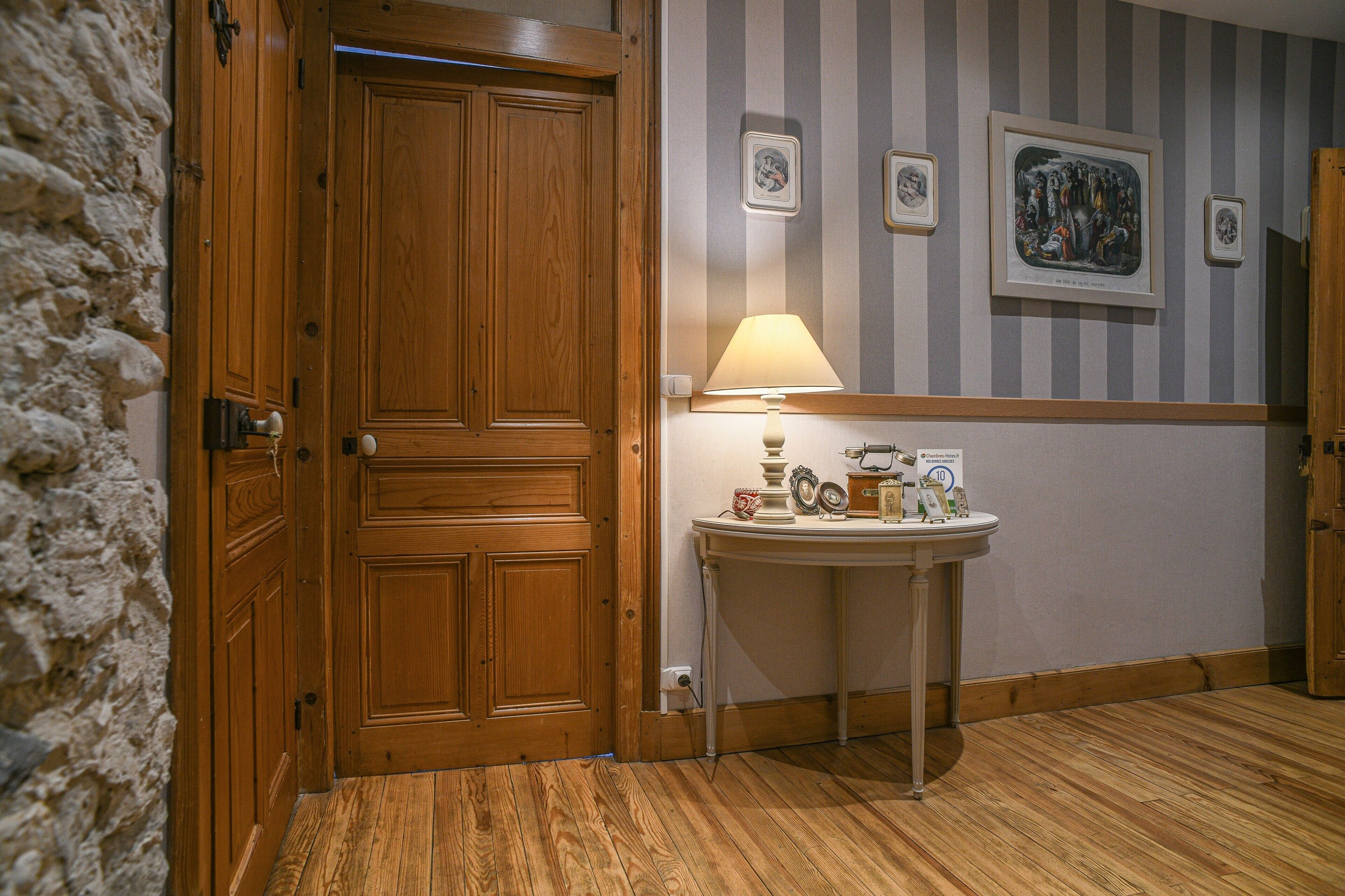 Ein Zimmer mit Holztür, einem kleinen Tisch mit Lampe und einer Wand mit gerahmten Bildern.