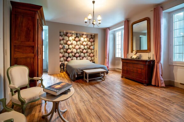 Une chambre à coucher comprenant un lit, une commode, une chaise, une table, un miroir et un mur recouvert d’un papier peint à motifs floraux.