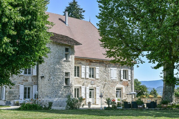Un bâtiment en pierre doté d’un toit en pente, de nombreuses fenêtres avec volets et d’un jardin bien entretenu avec des espaces pour s’asseoir à l’extérieur.