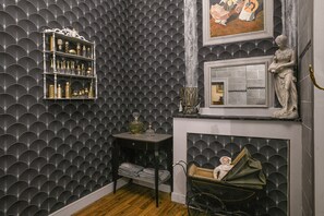 A room with a patterned wallpaper, a fireplace with a decorative mantel, and a small table with a lamp.