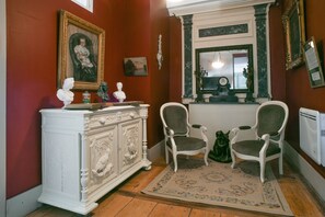 Une pièce comprenant une commode blanche ornée, deux fauteuils verts à motifs, un tapis à motifs et un miroir placé au-dessus d’une cheminée.