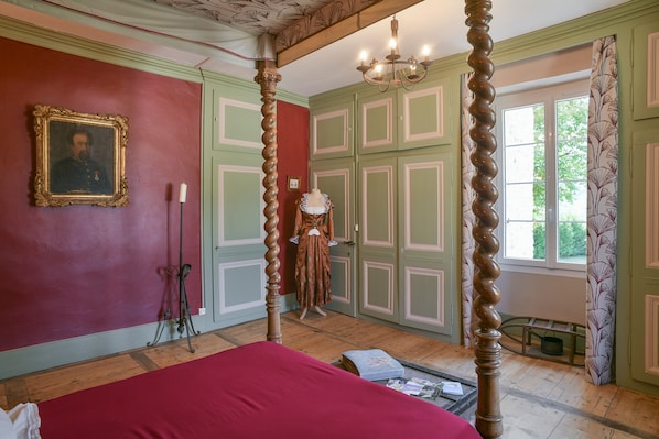 A room with a four-poster bed, a chandelier, a framed portrait on the wall, and a window with curtains.