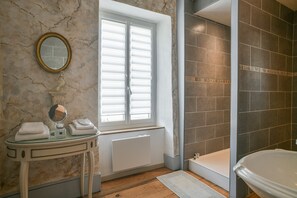 A bathroom with a marble wall, a window with blinds, a glass-topped vanity, a white bathtub, and a tiled shower area.