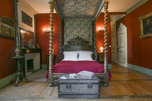 A four-poster bed with a red bedspread, a wooden chest of drawers, and a fireplace.