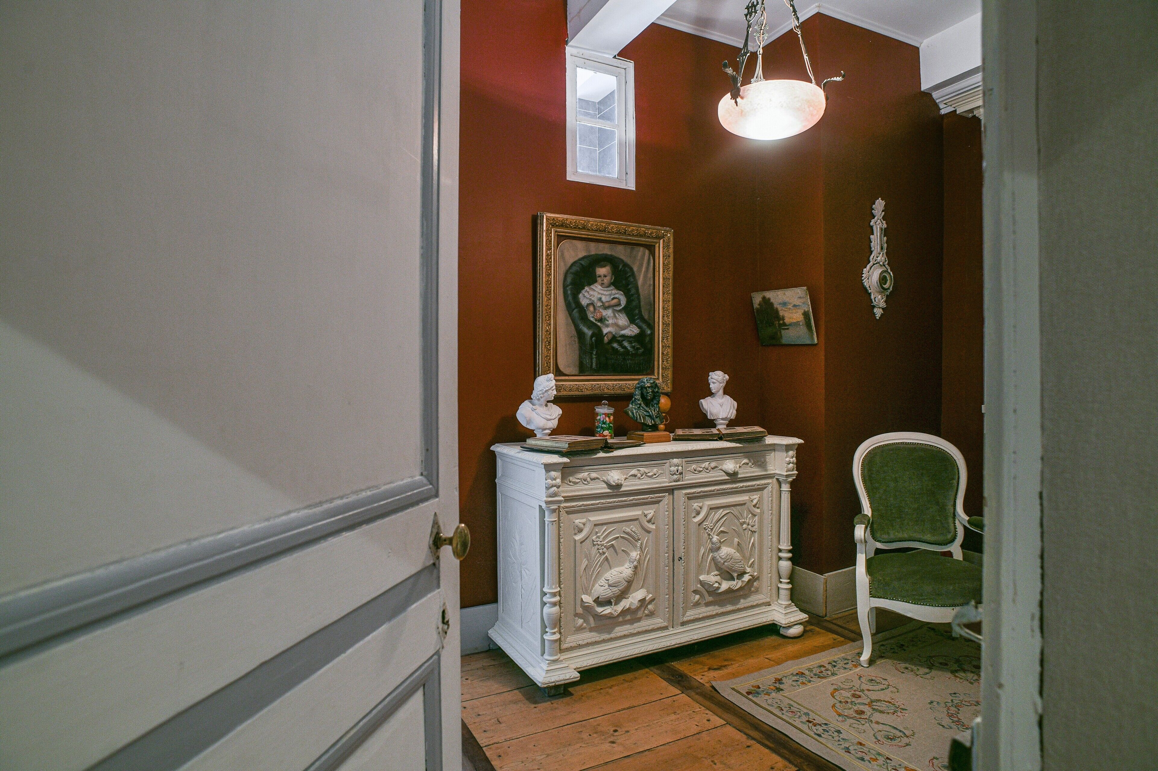 A hallway with a white ornate cabinet, a green upholstered chair, and a framed portrait on the wall.