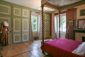 A room with a four-poster bed, green paneled walls, and a window with a view of greenery.