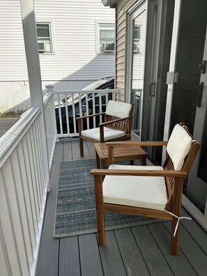 Eine Holzdielen-Terrasse mit zwei Stühlen und einem kleinen Tisch, einem blauen Teppich und einem weißen Gebäude im Hintergrund.