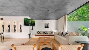Ein modernes Wohnzimmer mit Sofa, Couchtisch und Sesseln, einem Flachbildfernseher und einem großen Fenster mit Blick auf Grünflächen.