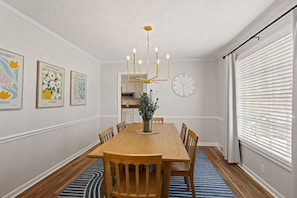 A dining room with a wooden table, chairs, a chandelier, wall-mounted clock, and framed floral artwork. - Pebble Creek Cottage (Taylors)