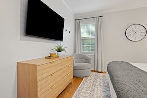 A bedroom with a bed, a wooden dresser, a grey armchair, a wall-mounted TV, a window with curtains, and a wall clock. - Pebble Creek Cottage (Taylors)