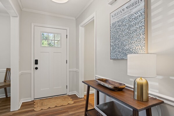 A hallway with a white door, a wooden bench, a table with a tray and a lamp, and a framed artwork on the wall.