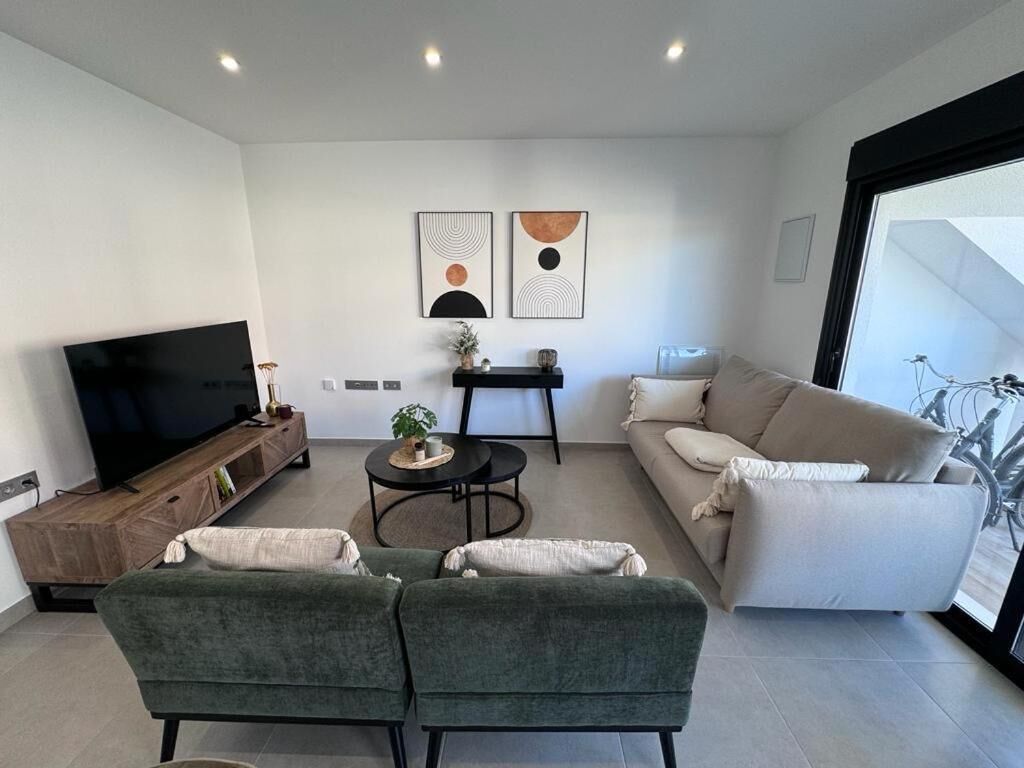 A modern living room with a flat-screen TV, two green sofas, a beige sofa, a small round table, and abstract wall art.
