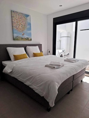 A modern bedroom with a large bed, a painting on the wall, and a view of the outdoors through a sliding door.
