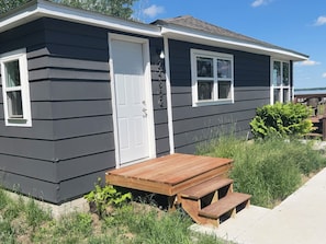 A single-story house with a dark exterior, white trim, a front door, two windows, a wooden deck, and a small garden area.