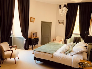 Ein Schlafzimmer mit einem Bett, einem Sessel, einer Kommode, einer Chandelüster und einem Fenster mit Vorhängen.