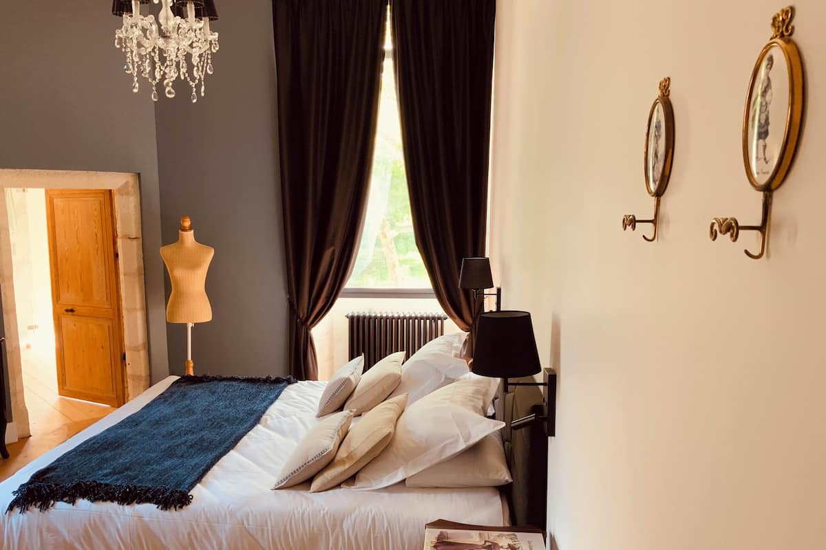 Ein Schlafzimmer mit einem Bett, einer Kommode, einer Lampe, einer Schönheitsmaske und einem Fenster mit Vorhängen.