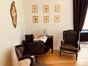 Una habitación con piso de madera, un sillón negro, una mesita auxiliar negra con tetera y tazas, y una pared decorada con impresiones botánicas enmarcadas.