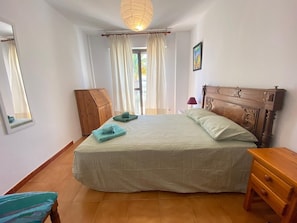 Een slaapkamer met een bed, een houten garderobe, een nachtkastje, een lamp, een spiegel en een raam met gordijnen.