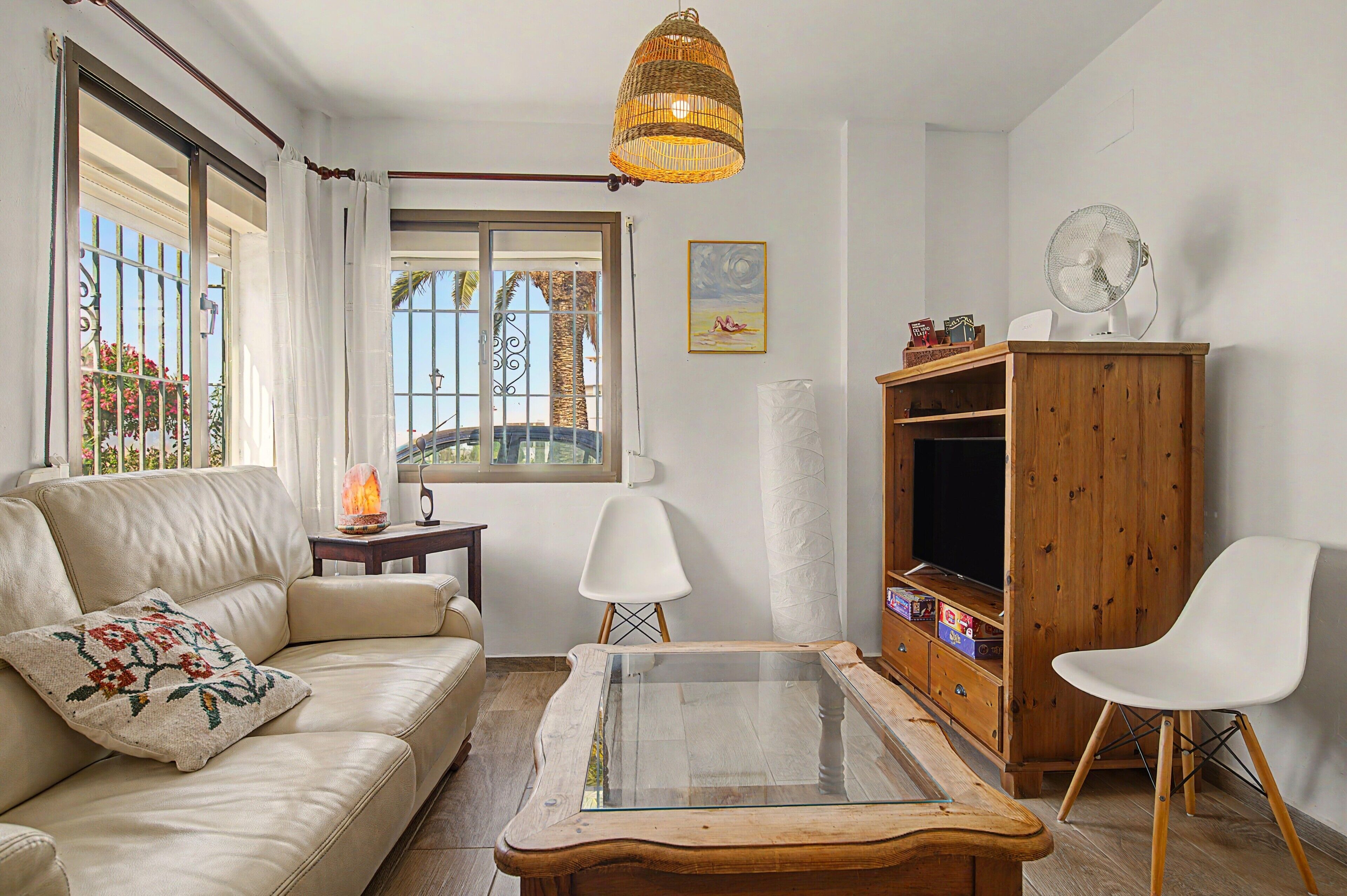 Una sala de estar con sofá, una mesa de centro de vidrio, un aparador de madera, una silla blanca, un ventilador y una ventana con vista a palmeras.