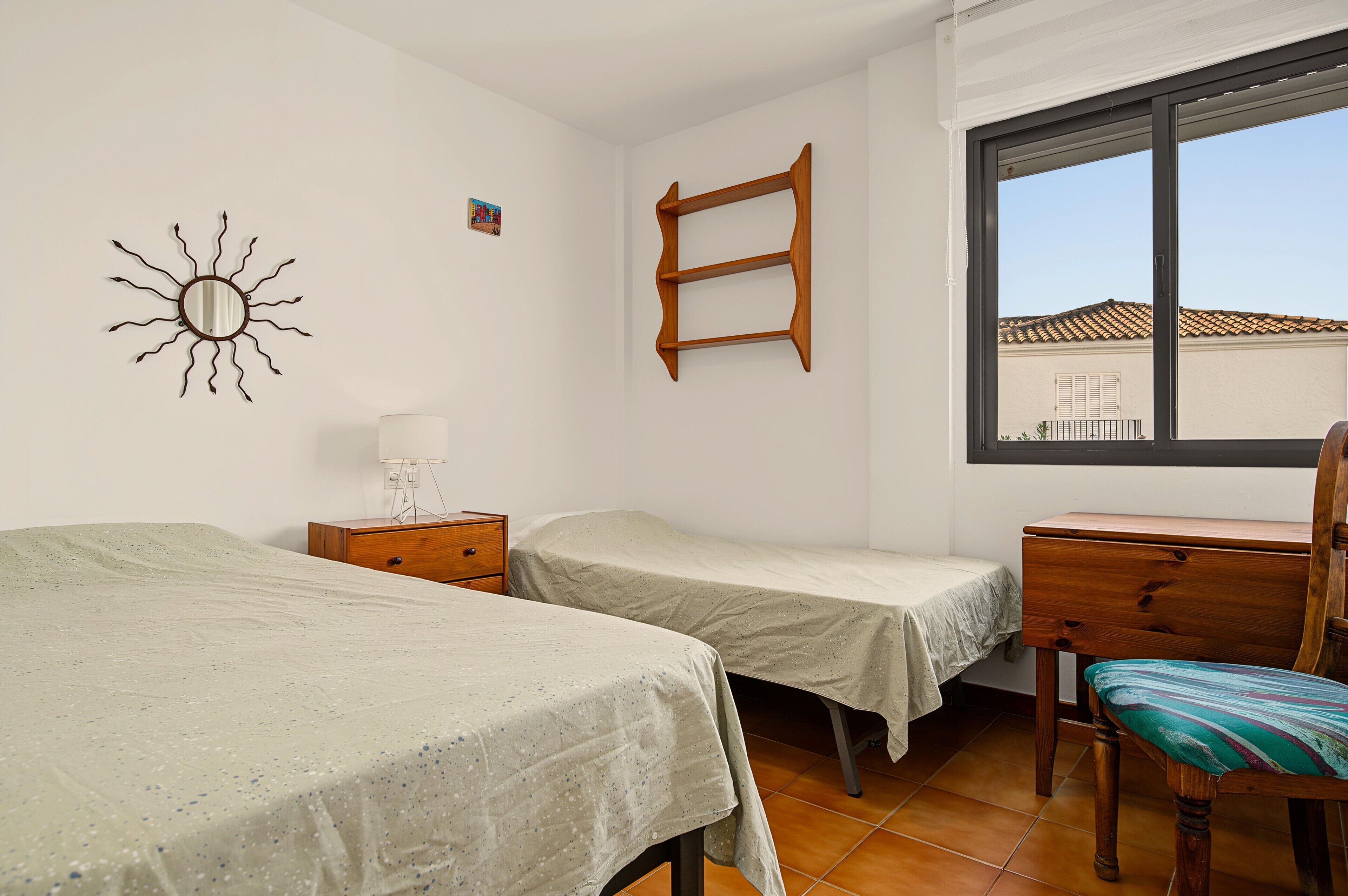 Un dormitorio con dos camas, una silla de madera, un escritorio, un ventanal y un estante en la pared.