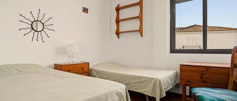 Un dormitorio con dos camas, una silla de madera, un escritorio, un ventanal y un estante en la pared.