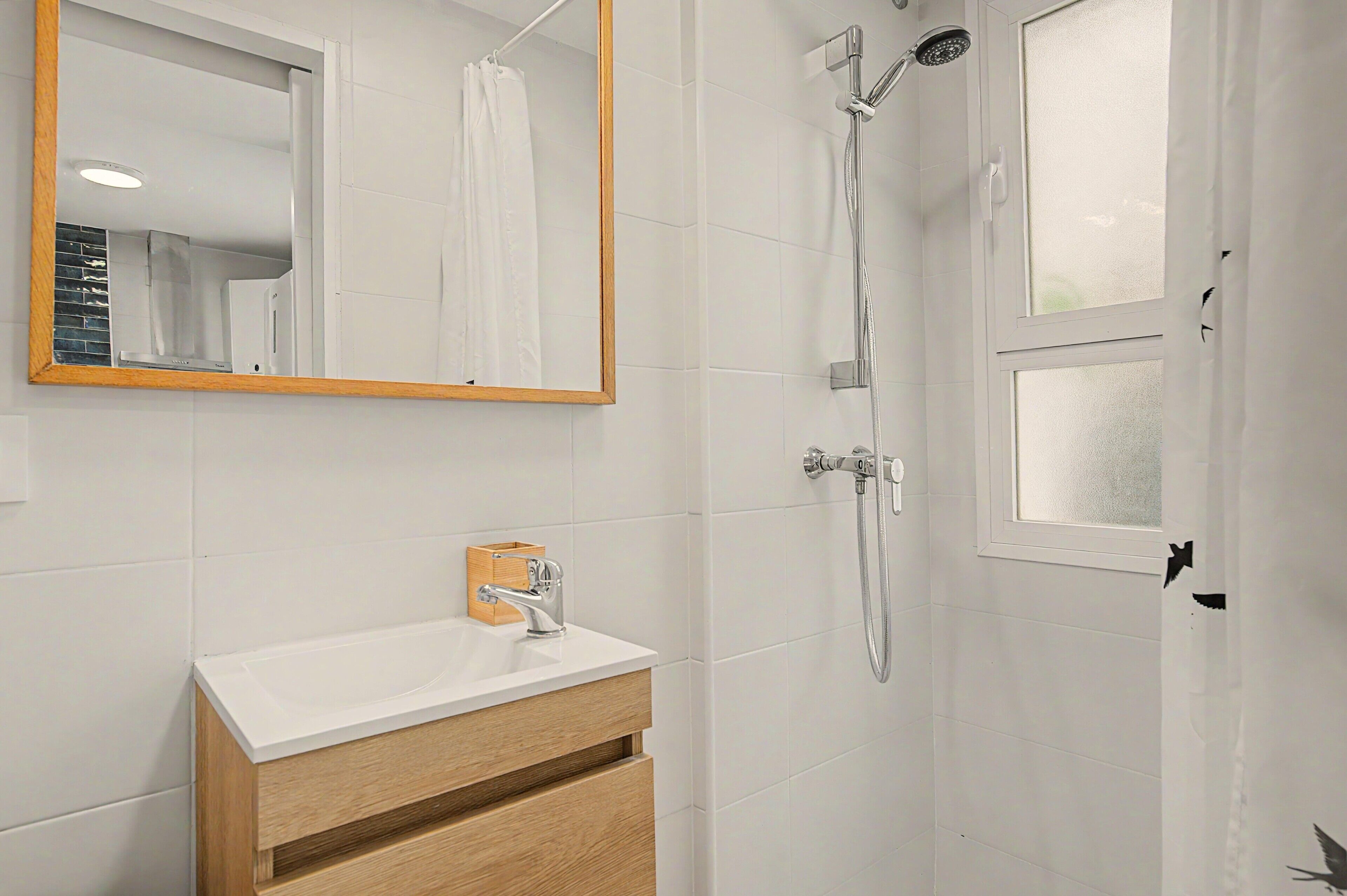 A bathroom with a white vanity, a mirror with a wooden frame, a shower with a handheld showerhead, and a window with a frosted glass panel.