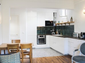 Eine moderne Küche mit weißen Schränken, einer Mikrowelle, einem Backofen und einer Kaffeemaschine. Es gibt einen Esstisch mit Stühlen und einen kleinen Teppich.