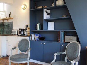 Ein Zimmer mit einem blauen Schrank, zwei grau gepolsterten Sesseln, einem kleinen Tisch mit einem Wasserkocher und einem Regal mit Büchern und Dekorationsobjekten.
