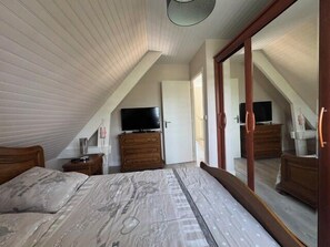 Ein Schlafzimmer mit schrägem Dach, einem Bett, einer Kommode, einem Fernseher und einem großen Spiegel.