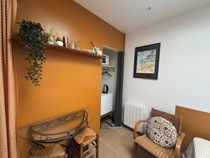 A room with a bed, a chair, a small table, a radiator, a shelf with plants, and a picture on the wall. - Charming studio apartment with garden in Ifs (Ifs)