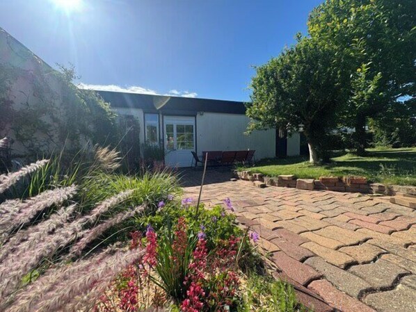 A building with a paved patio, a tree, and flowering plants. - Charming studio apartment with garden in Ifs (Ifs)