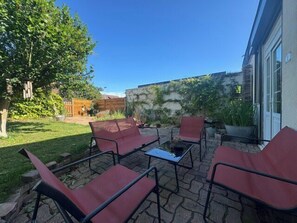 An outdoor patio with four red chairs and a table, a tree, and a building in the background. - Charming studio apartment with garden in Ifs (Ifs)