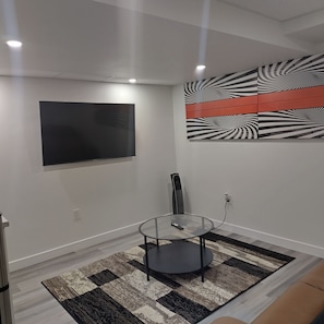 A modern living room with a flat-screen TV, a glass table, a standing speaker, and a patterned rug.