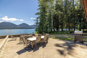 Un patio exterior con mesa y sillas, una parrilla y vistas a un lago y montañas.