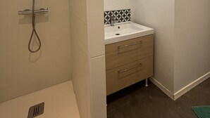 Une salle de bain avec une douche, un lavabo, un miroir et un mur recouvert de carreaux derrière le lavabo.