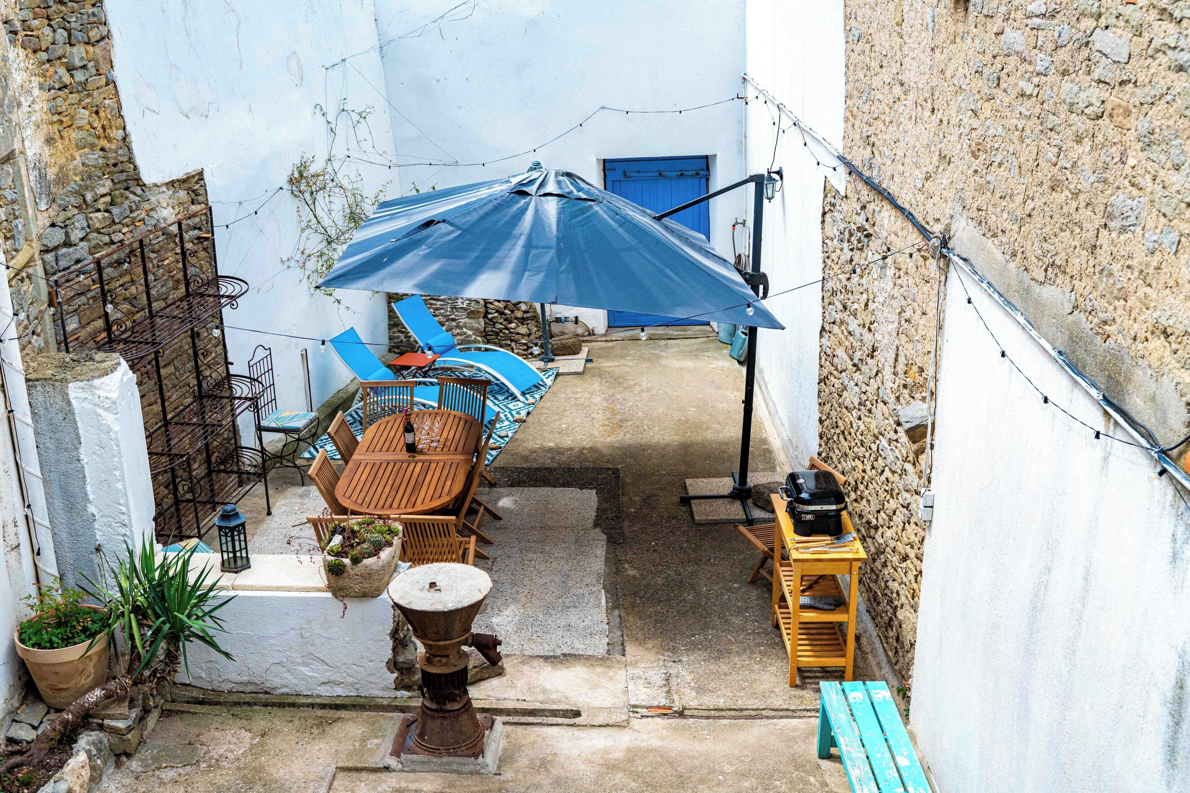 A courtyard with a table and chairs, a blue umbrella, a wooden bench, and a stone wall.