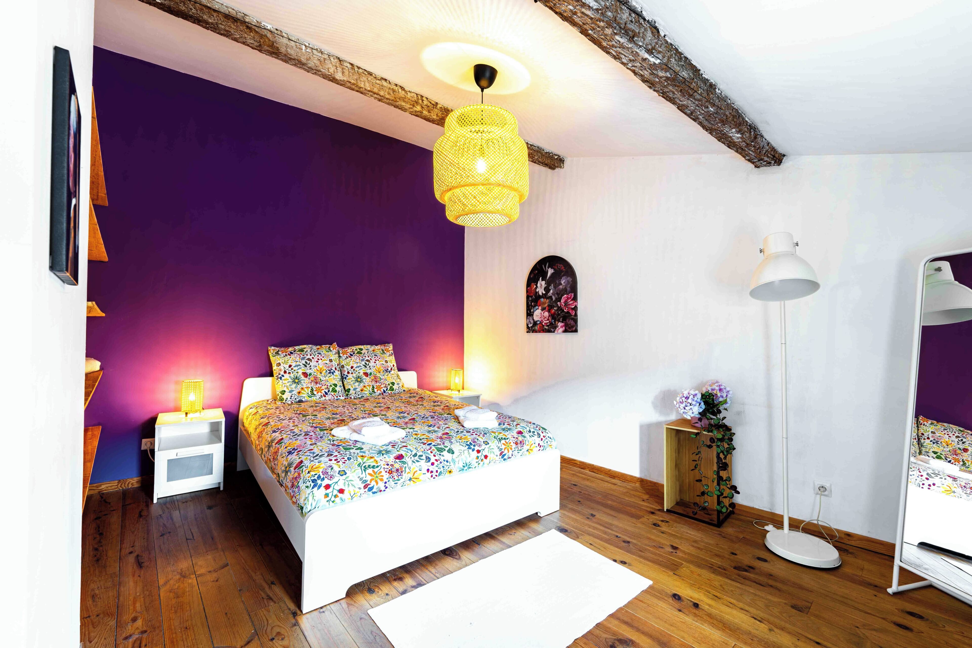 Une chambre avec un plancher en bois, un lit recouvert d’une literie à motifs floraux, un luminaire suspendu et une petite table de chevet avec une lampe.