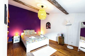A bedroom with a wooden floor, a bed with floral bedding, a hanging light fixture, and a small bedside table with a lamp. - L'Escapade Minervoise, perfect for families or friends   (Laure-Minervois)
