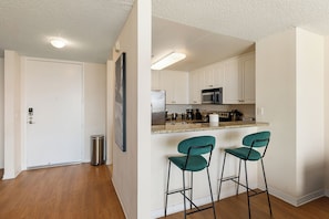 A kitchen with white cabinets, a stainless steel refrigerator, a microwave, and a sink. There are two green bar stools and a trash can. - Sunset Pearl' Full Ocean Views Pool Gym & Parking Available (Marina del Rey)