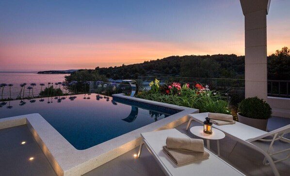 Ein Infinity-Pool mit Meerblick bei Sonnenuntergang, Sitzgelegenheiten im Freien und ein Topfpflanze.
