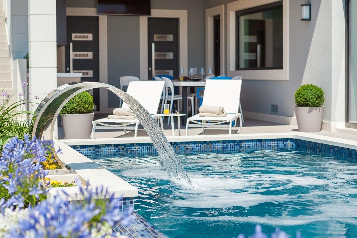 A swimming pool with a water feature, surrounded by white lounge chairs and a table with chairs, set against a modern building facade.