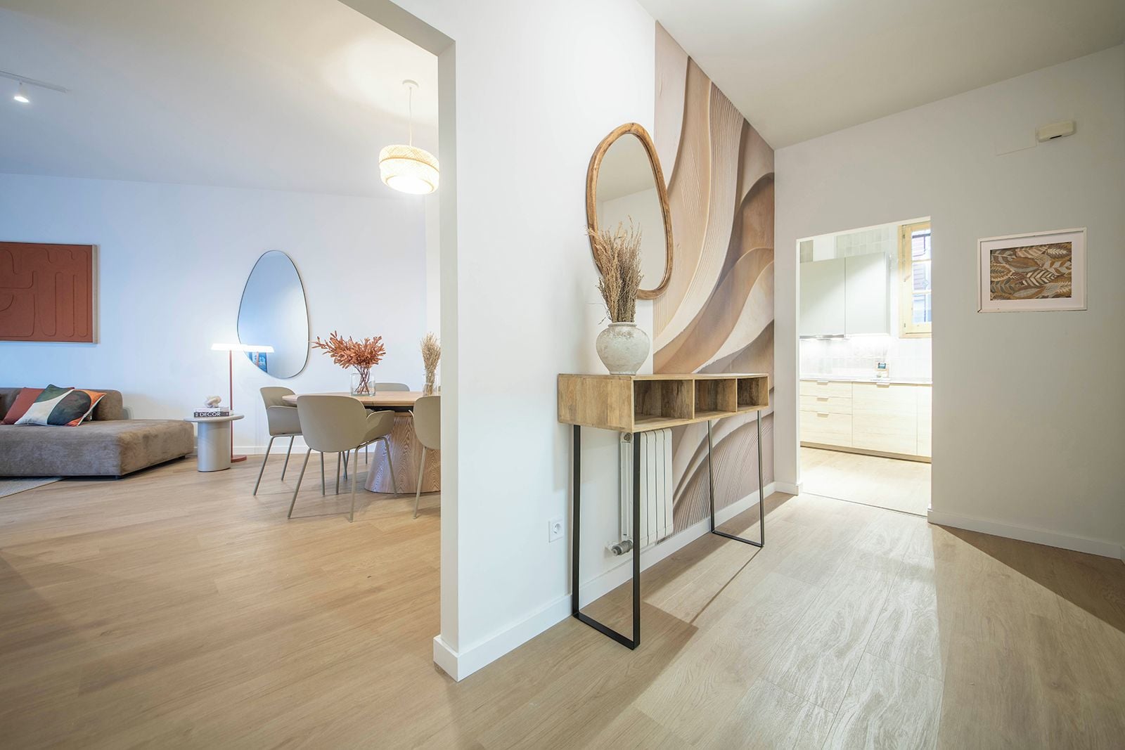 A modern living room with a sofa, a dining table, and chairs. There is a console table with a vase and a mirror. The room has wooden flooring and a large window.