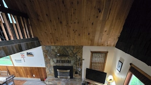 A cozy living room with a stone fireplace, a flat-screen TV, and a large window with a view of the outdoors. - ~ Riverfront, Hot Tub, Mountain Views, 8 Beds ~ (Cascade)