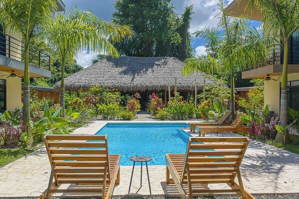 A pool with wooden chairs, a thatched-roof structure, and tropical plants. - Walk to Beach, 4 BR/ 4BA Villa, Pool Garden Rancho (Playa Grande)