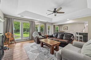 Ein geräumiges Wohnzimmer mit Kamin, Ledersofas, einem Holztisch und großen Fenstern mit Vorhängen.