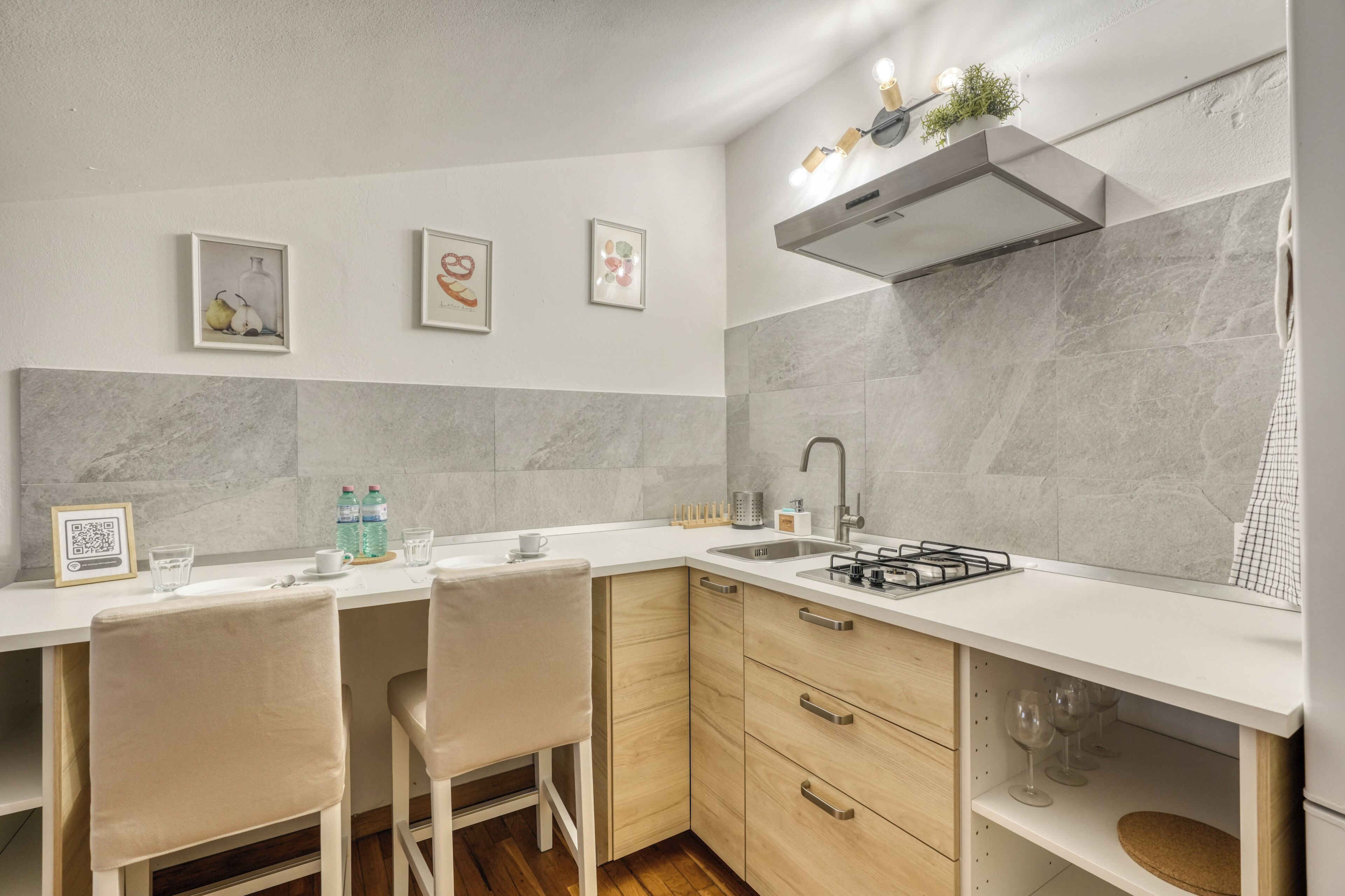 A compact kitchen with a white countertop, two bar stools, a built-in oven, and a stainless steel sink.