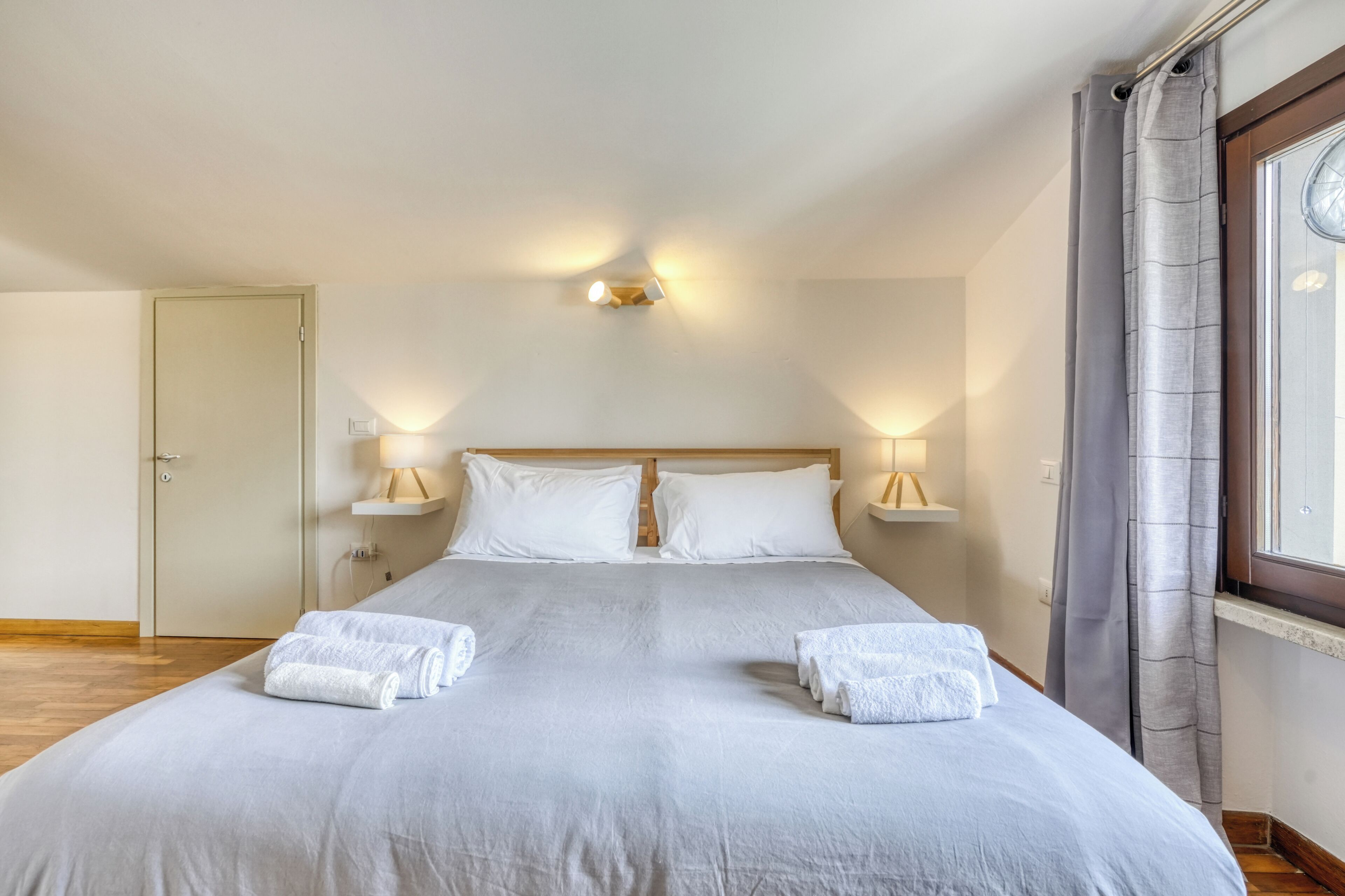 Una cama bien hecha con sábanas y toallas blancas, dos lámparas de noche, un cabecero de madera y una ventana con cortinas.