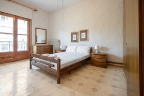 A bedroom with a wooden bed, two bedside tables with lamps, a dresser, a large window, and two framed pictures on the wall.